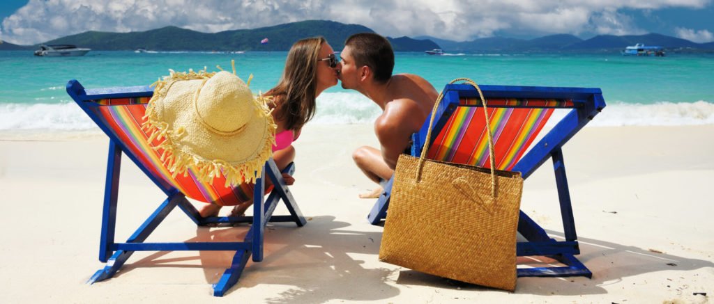 a man and a woman kissing on a beach with chairs and water in the background